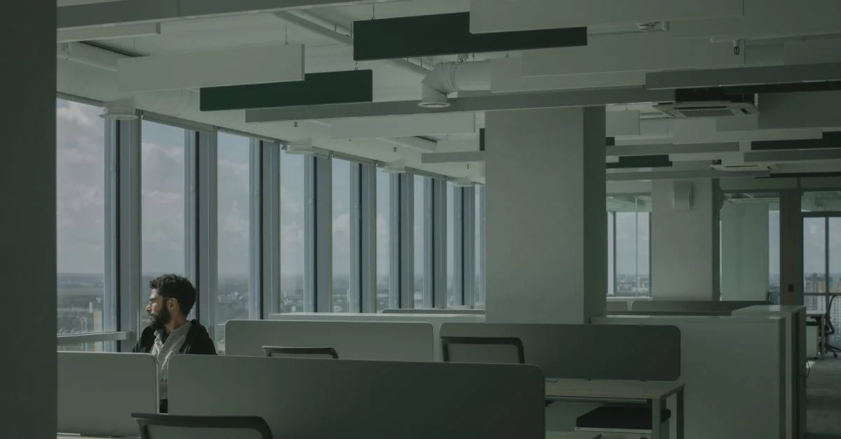A man sits in an open-plan office with tall windows and partitioned workstations.